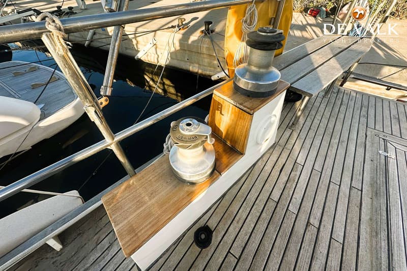 Twee Lewmar lieren, een houten dek en een reling zijn zichtbaar op de boot, met water en een kade op de achtergrond.