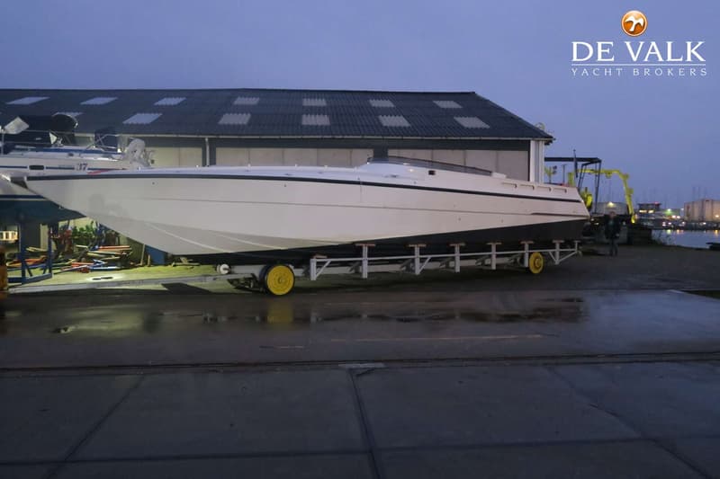 Een witte speedboot met een zwarte streep staat op een trailer met gele wielen voor een gebouw met een donker dak.