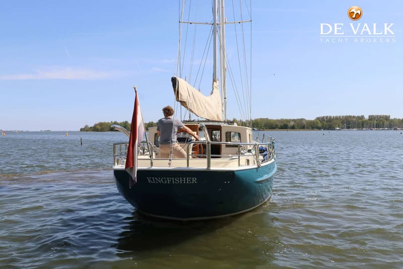Een zeilboot met de naam "KINGFISHER" vaart op een meer met een persoon aan het roer en een Nederlandse vlag.
