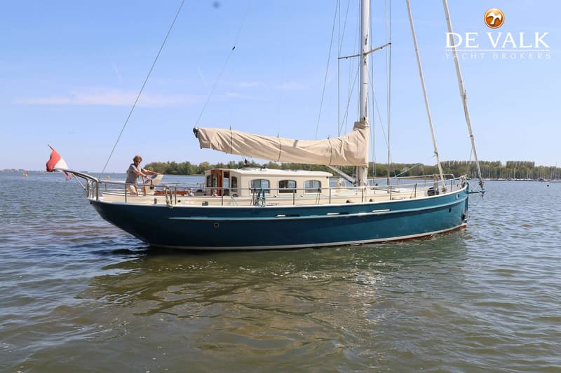 Een blauwe zeilboot met een beige kajuit en opgerold zeil vaart op het water, met een persoon aan het roer.