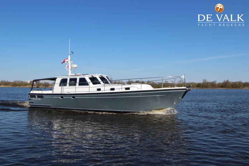 Een grijze motorboot met witte opbouw vaart op een blauw water met een blauwe lucht en bomen op de achtergrond.