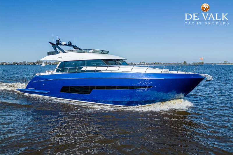 Een blauw-witte motorboot vaart over het water, met een logo van "DE VALK YACHT BROKERS" in de rechterbovenhoek.