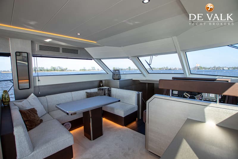 De foto toont het interieur van een boot met een zithoek, tafel, stuurstand en uitzicht op water en landschap.