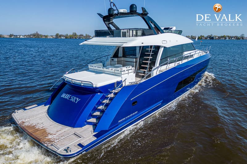 Een blauw-witte motorjacht met de naam "BLUE SKY" vaart op een waterweg met bebouwing op de achtergrond.