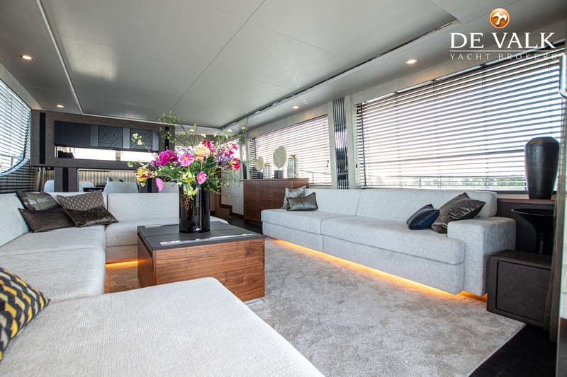 De foto toont een modern interieur met twee lichte banken, een houten salontafel met bloemen en ramen met jaloezieën.