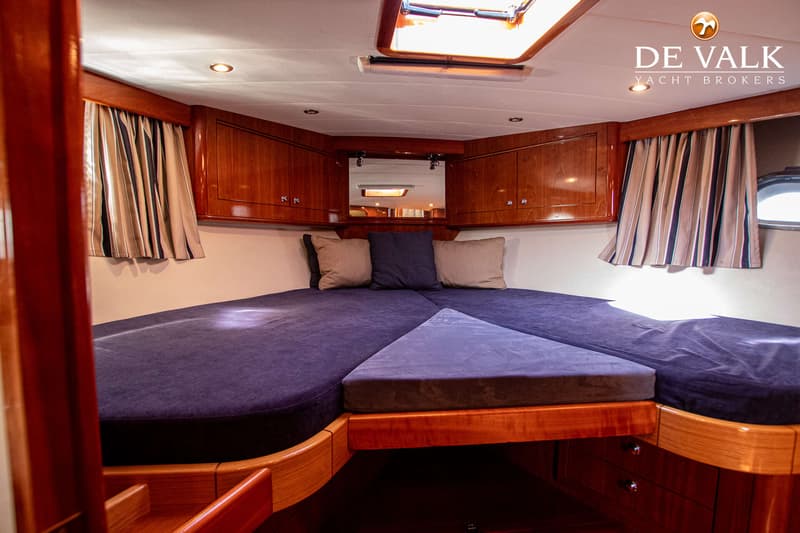 De foto toont een kajuit met een V-vormig bed met donkerblauwe kussens, omringd door houten kasten en gordijnen.