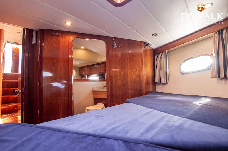 De foto toont een kajuit met twee bedden, een houten interieur, een patrijspoort met gordijnen en een badkamer met toilet.