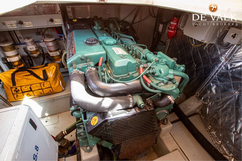 Een groene scheepsmotor met slangen en bedrading, een gele tas, een brandblusser en een bedieningspaneel zijn zichtbaar.