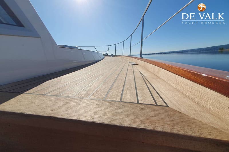 Een houten dek van een boot met een reling, een witte opbouw en een glimmende bruine rand, met water en land op de…