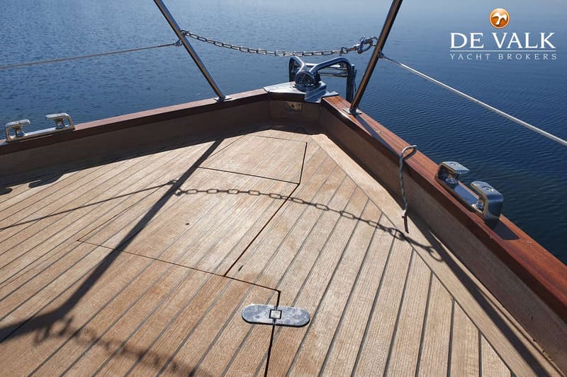 Een houten dek met een ankerlier, reling en ketting aan de boeg van een boot op het water. — Martin 44 Trawler