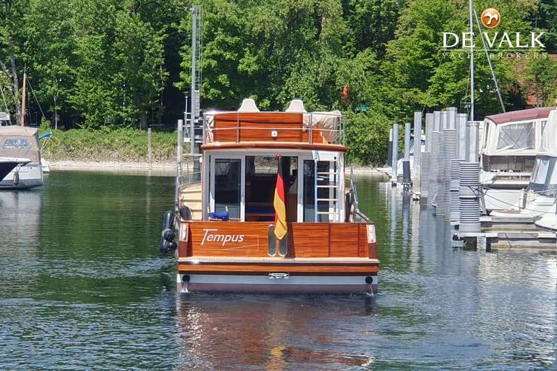 Een houten boot met de naam "Tempus" en een Duitse vlag vaart op het water, omringd door bomen en andere boten.