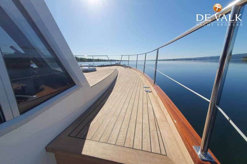 De foto toont het voordek van een boot met houten planken en een metalen reling, varend op rustig water onder een heldere…