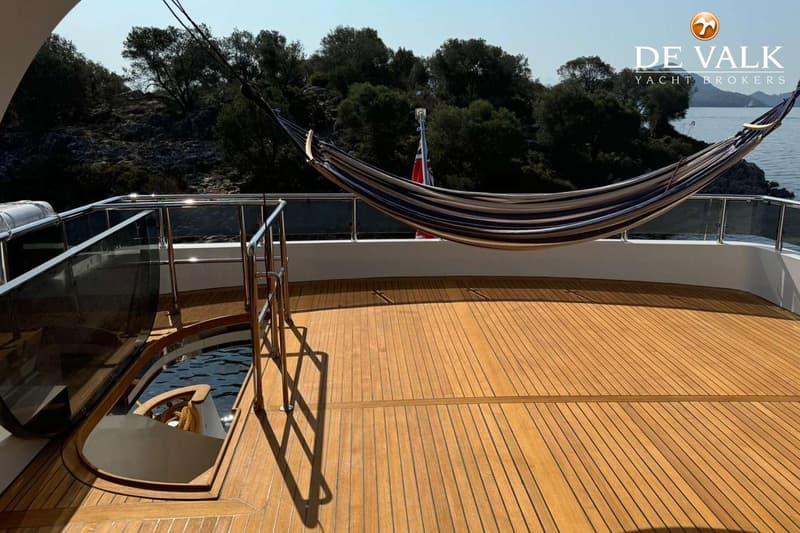 Een houten dek met een hangmat, een glazen reling en een opening naar een lager gelegen gedeelte met water.