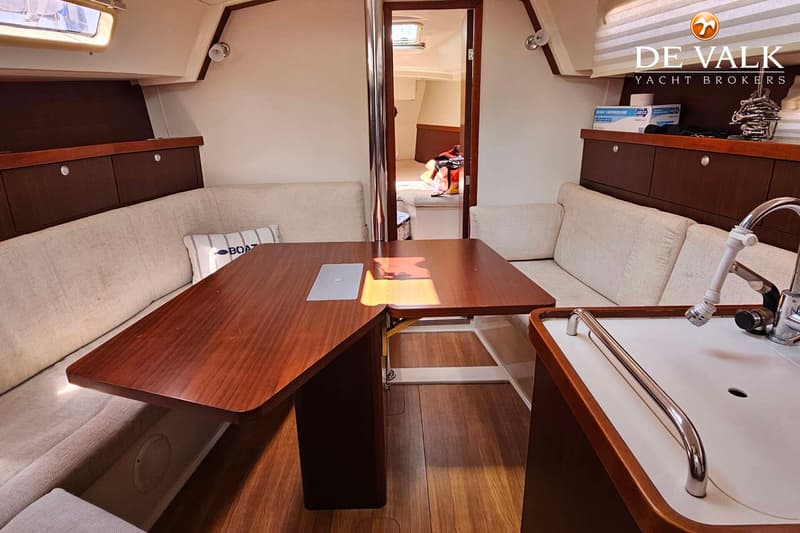 Een interieur van een boot met een houten tafel, banken met lichte kussens, een gootsteen en een doorgang naar een andere…
