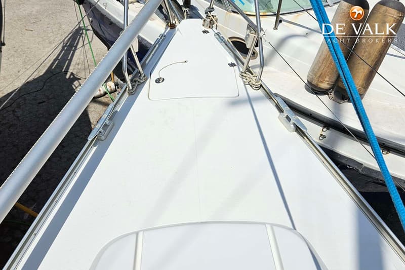 De foto toont het witte dek van een boot met een luik, reling en touwen, met op de achtergrond een andere boot en twee…