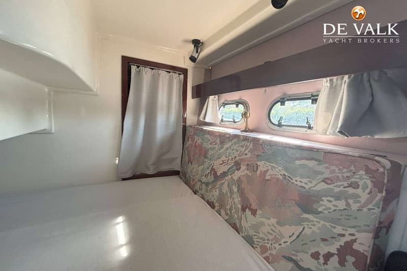 Een kajuit met een bed, twee patrijspoorten met gordijnen, een leeslampje en een kussen met een camouflagepatroon.