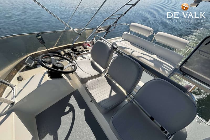 Een bovenaanzicht van de cockpit van een boot met een stuurwiel, instrumentenpaneel, grijze stoelen en een bankje.
