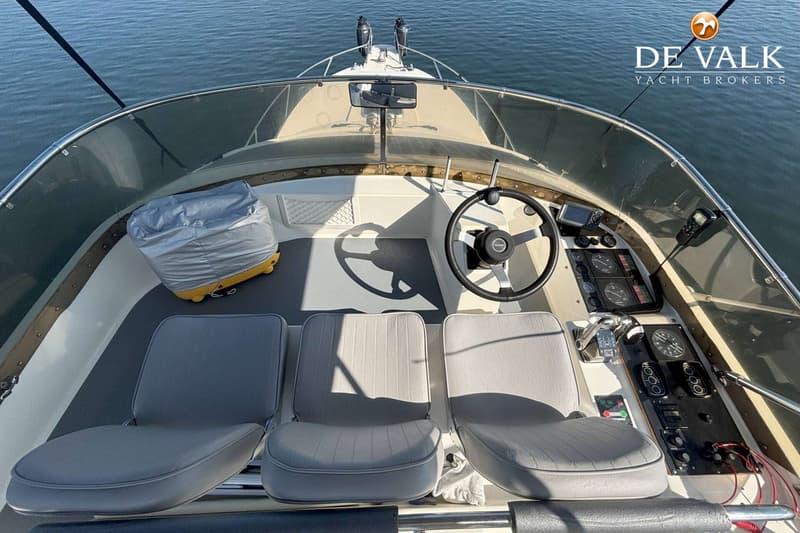 Een bovenaanzicht van de cockpit van een boot met een stuurwiel, instrumentenpaneel, drie stoelen en een afgedekte koelbox.