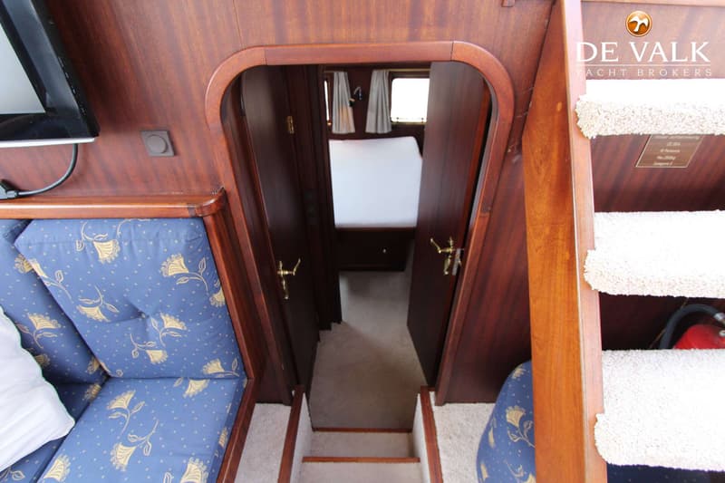 Een interieur van een boot met houten wanden, een blauwe bank met bloemmotief, een trap en een doorgang naar een slaapcabine.