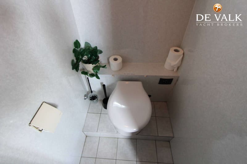 Een witte toiletpot met een plantje, toiletpapier en een toiletborstel in een kleine ruimte met betegelde vloer.