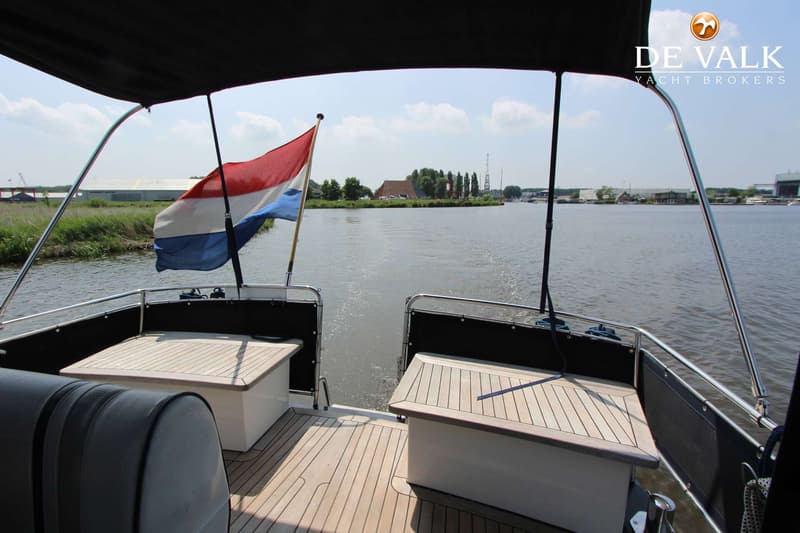 De foto toont de achterkant van een boot met een Nederlandse vlag, twee houten banken en een waterweg met landschap op de…