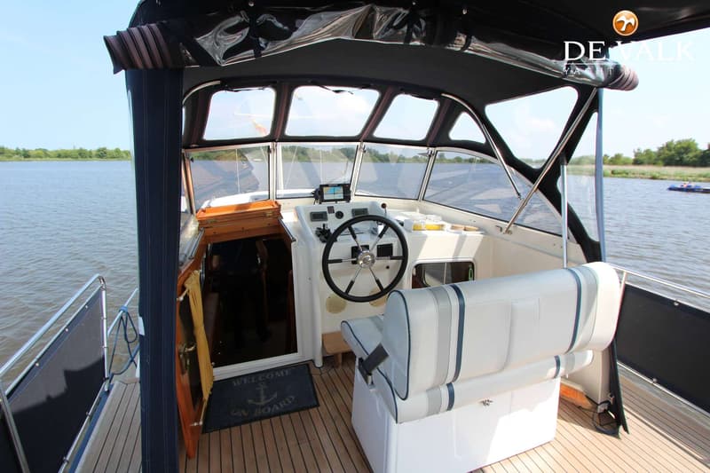 Een bootinterieur met stuurwiel, navigatieapparatuur, witte banken en een houten vloer, met uitzicht op water.
