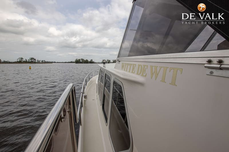 Een witte boot met de naam "WITTE DE WIT" in gouden letters vaart op een meer met een gele boei en bomen op de achtergrond.