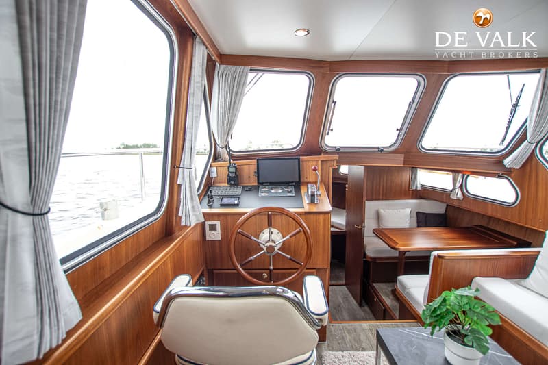 De foto toont het interieur van een boot met een stuurstand, houten afwerking, zitbanken en ramen met uitzicht op water.