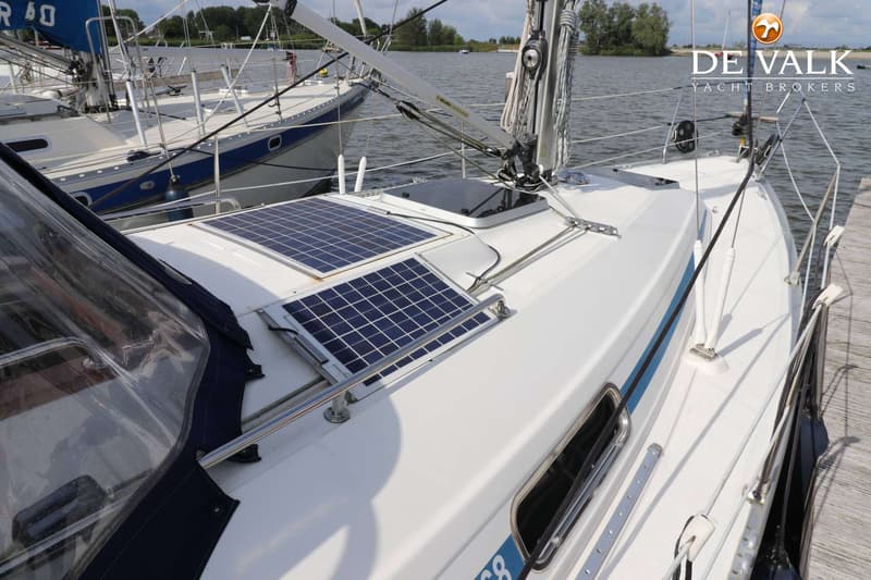 De foto toont het dek van een witte zeilboot met twee zonnepanelen en een luik, met op de achtergrond water en een andere…