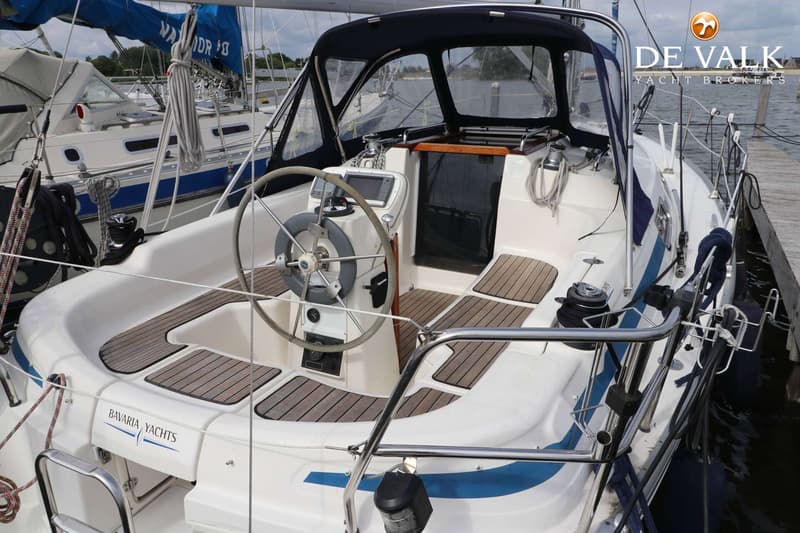 Een witte zeilboot met blauwe accenten en een donkerblauwe buiskap, met een stuurwiel en houten dekdelen. — Bavaria 31