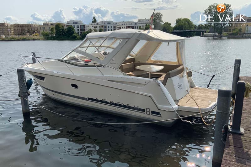 Een witte motorboot met een grijze kap ligt aangemeerd aan een houten steiger, met gebouwen op de achtergrond.