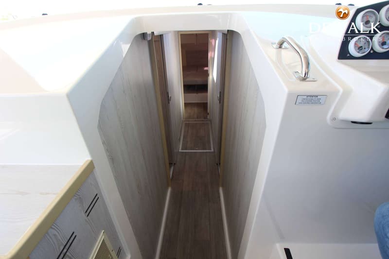 Een smalle gang met houten vloer en lichtgekleurde wanden leidt naar een slaapruimte, met een stuurconsole rechts.