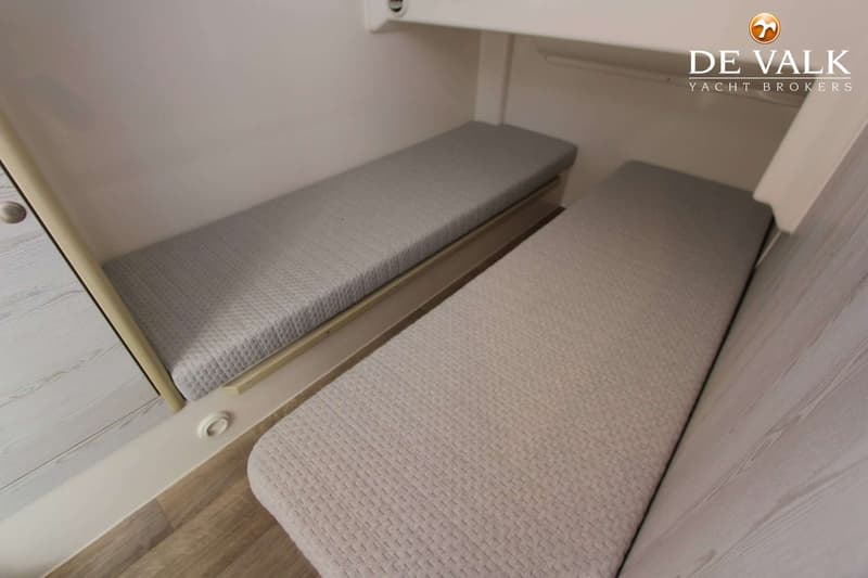 Twee smalle bedden met grijze matrassen zijn zichtbaar in een kajuit met lichte wanden en een houten vloer.