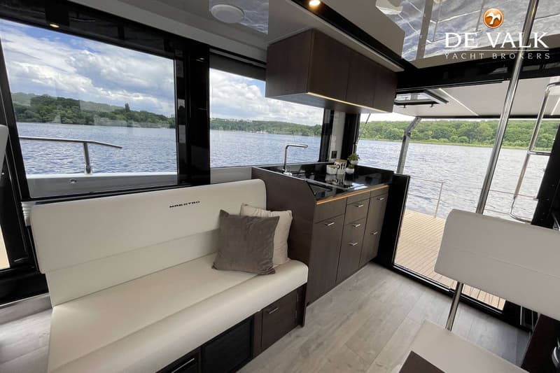 Een interieur van een boot met een witte bank, een donkere keuken en grote ramen met uitzicht op water en beboste oevers.