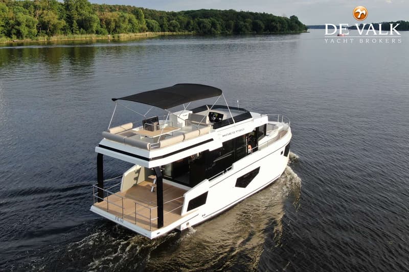 Een witte motorboot met een zwart dak en een houten achterdek vaart op een meer met bosrijke oevers.