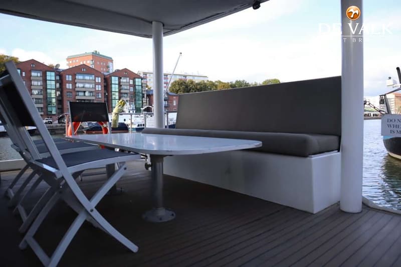 Een bootdek met een witte tafel, twee opvouwbare stoelen, een vaste bank en een uitzicht op gebouwen en water.