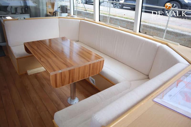 Een interieur van een boot met een houten tafel en een U-vormige bank met lichte kussens, omringd door ramen.