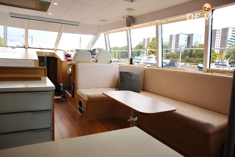 De foto toont het interieur van een boot met een stuurstand, zitbanken, een tafel en ramen met uitzicht op gebouwen en…