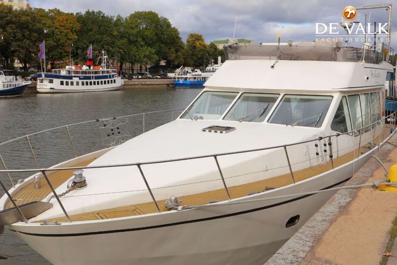Een witte motorboot met een houten dek ligt aangemeerd aan een kade, met andere boten en bomen op de achtergrond.