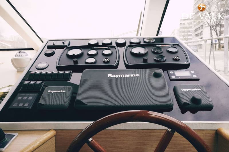 De foto toont een stuurstand met een houten stuurwiel, diverse meters, schakelaars en Raymarine apparatuur.