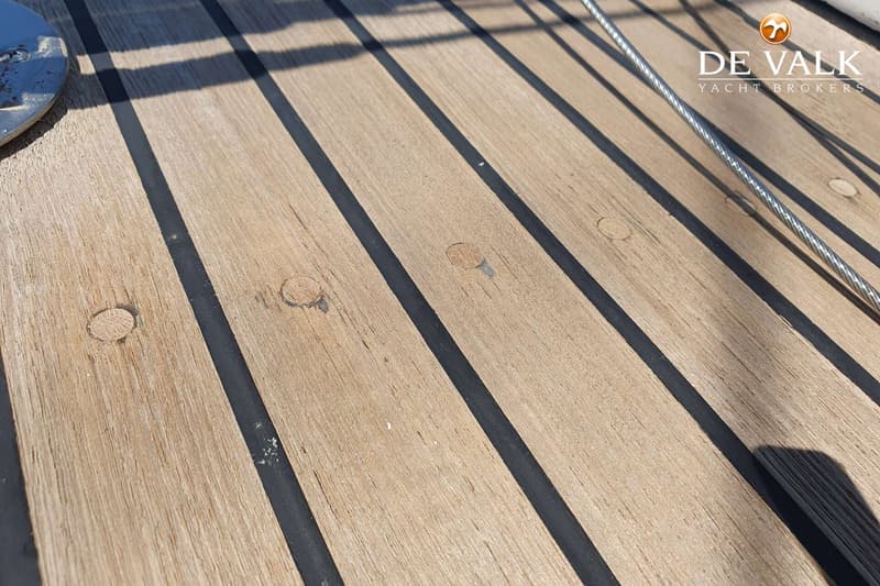 Een close-up van een houten scheepsdek met zwarte naden, een metalen plaat en een staalkabel.