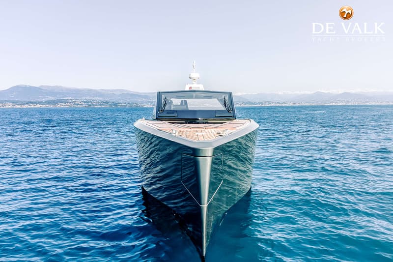 Een donkergrijze motorboot met een houten dek en een zwarte voorruit ligt op blauw water met bergen op de achtergrond.