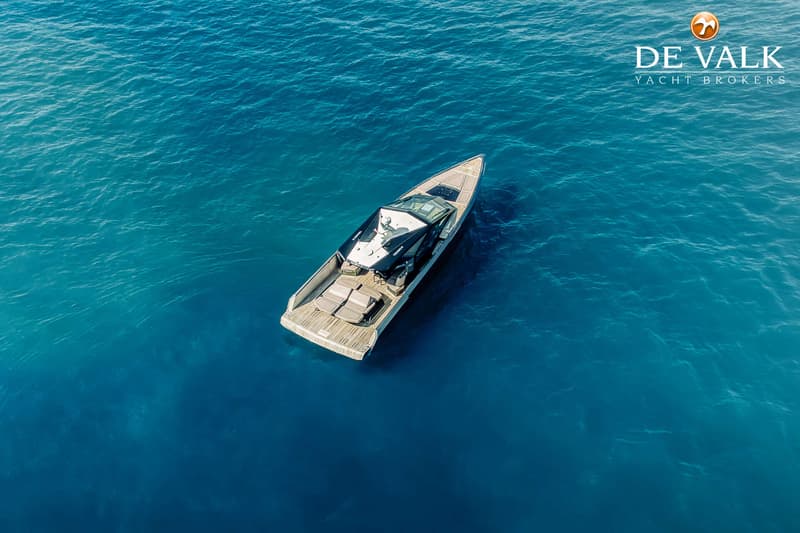 Een motorjacht met een houten dek en een zwarte kap ligt in helderblauw water, met een logo van "DE VALK YACHT BROKERS"…