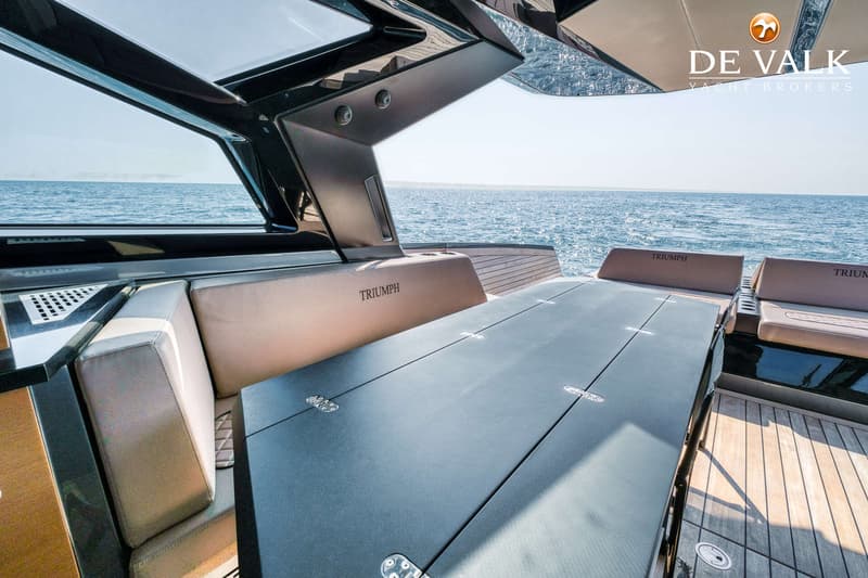 Een bootinterieur met beige kussens en een donkere tafel, met de zee zichtbaar op de achtergrond.