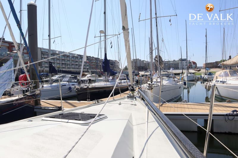 Een bovenaanzicht van het dek van een witte zeilboot met een luik, touwen en reling, omringd door andere boten in een haven.