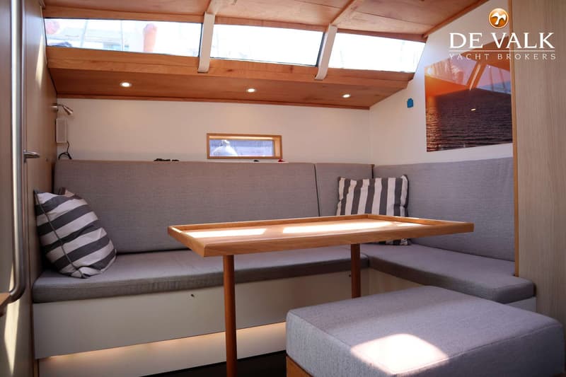 Een interieur van een boot met een houten tafel, grijze banken en kussens, en ramen in het plafond en de wand.