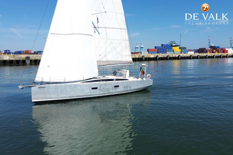Een witte zeilboot met een Belgische vlag vaart op het water, met een haven met containers op de achtergrond.