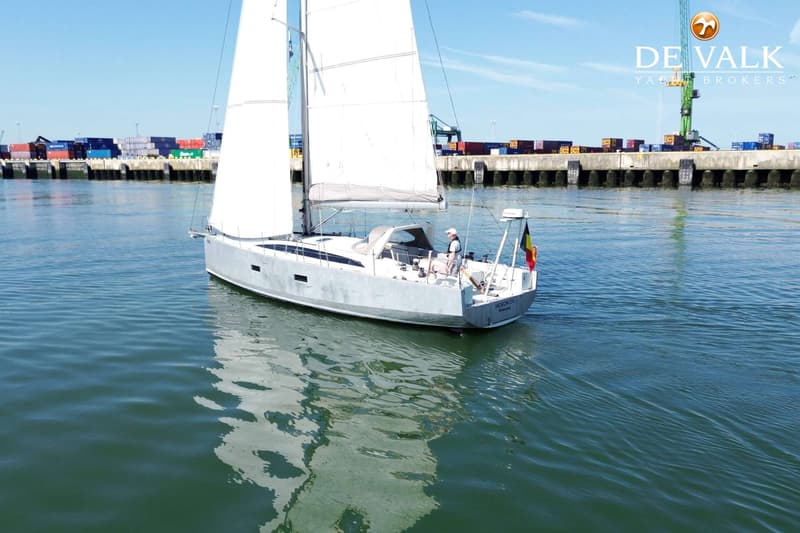 Een witte zeilboot met een Belgische vlag vaart op rustig water, met een haven vol containers op de achtergrond.