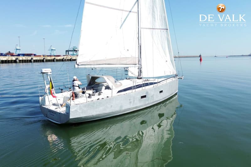 Een zeilboot met de naam "ANDROMEDA Antwerpen" vaart op rustig water, met een persoon aan het roer en een haven op de…