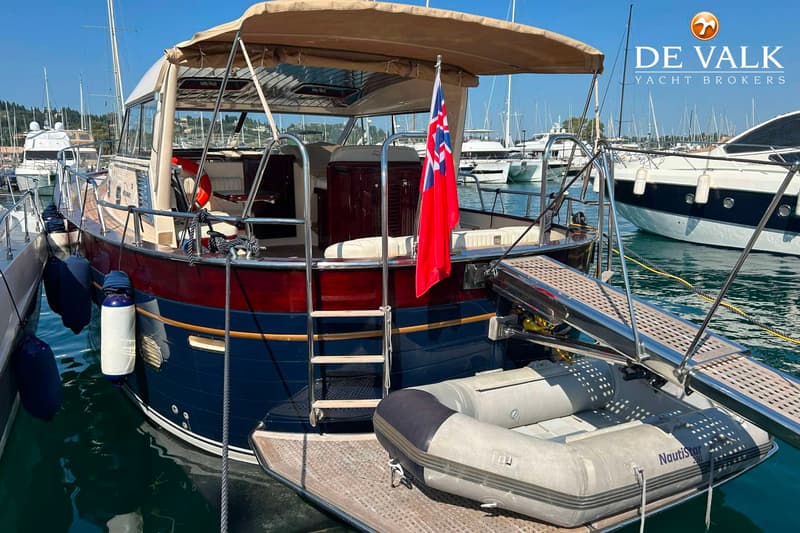 Een donkerblauwe boot met een rood-bruine opbouw en een beige zonnedek ligt in een haven met een bijboot op het zwemplateau.